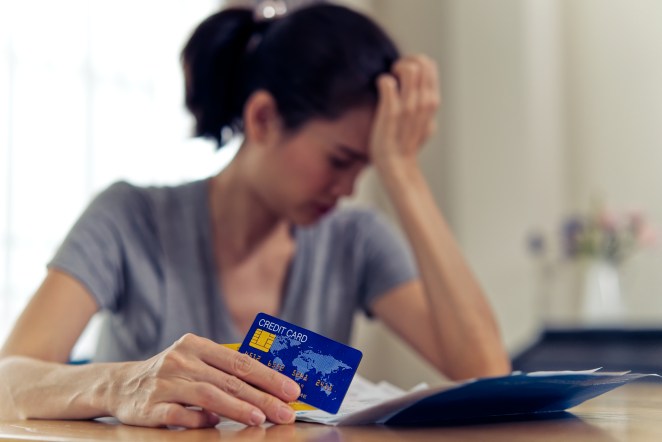 Stressed young sitting Asian woman hands holding credit card and bills worry about find money to pay credit card debt and all loan bills. She is putting hand on her head. Financial problem concept.